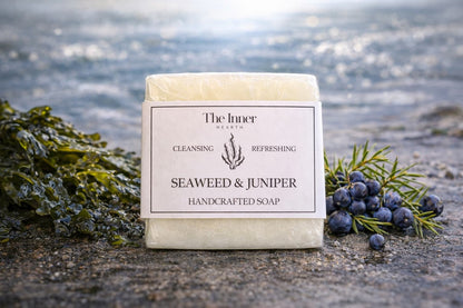Handcrafted soap bar with 'Seaweed & Juniper' scent on a beach with seaweed and juniper berries.